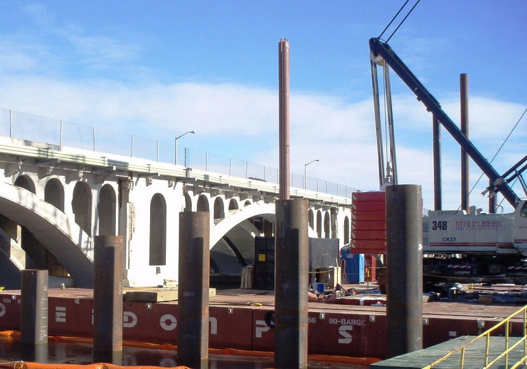 The construction of the Kenneth F. Burns Memorial Bridge was a complex and high-profile project that replaced a structurally deficient, 7-span concrete arch bridge built in 1916 with 2 new bridges over Lake Quinsigamond, between Worcester and Shrewsbury, MA. The new bridges are 870’ long, 5-span open spandrel steel deck arches founded on 36” diameter pipe piles up to 290’ deep. The project also included reconstructed portions of Lake Avenue and Route 9 and contains many architectural features.